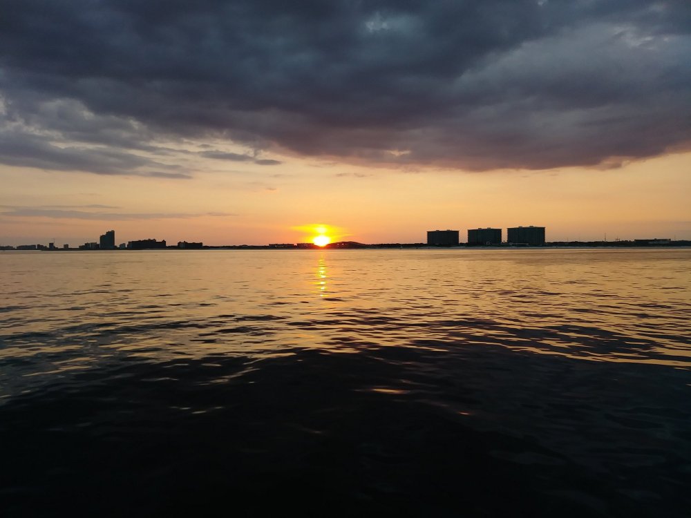 Sunset over calm water with silhouettes of buildings and cloudy sky.