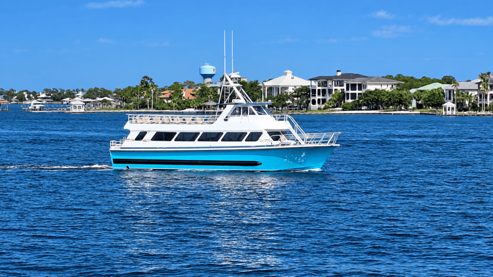 A blue and white boat cruises on a sunny lake with houses and trees in the background.